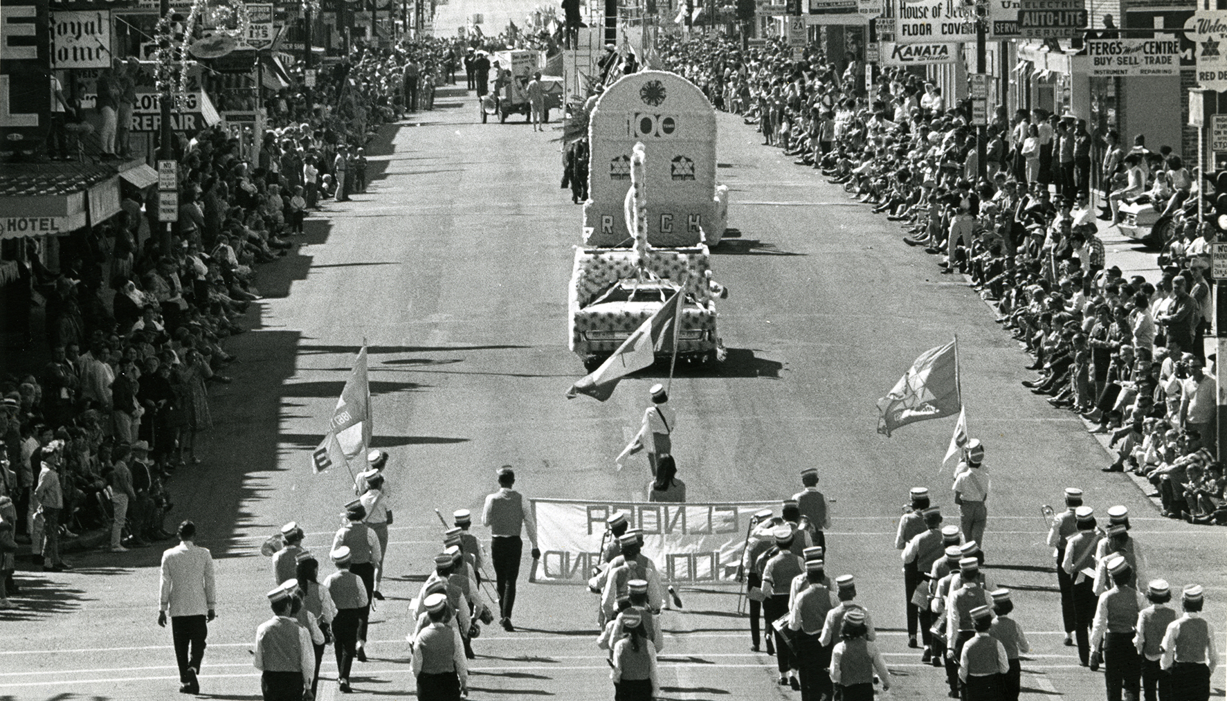 Black and white photo of a large crowd sitting on both sides of a wide street watching the Elnora Marching Band in a parade