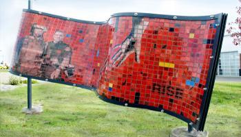 Red side of a rectangle sculpture made up of glass tiles an the photo etched tiles that show police and emergency services personnel and and out-reading hands trying to join. The word 